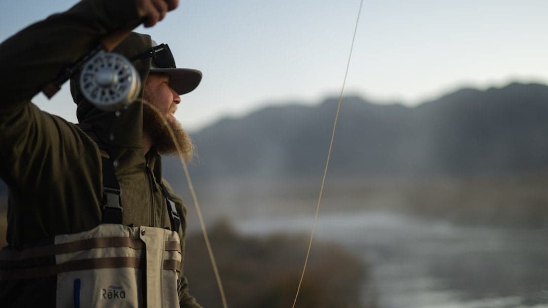 Fly angler casting at sunrise on a calm river with mountain backdrop