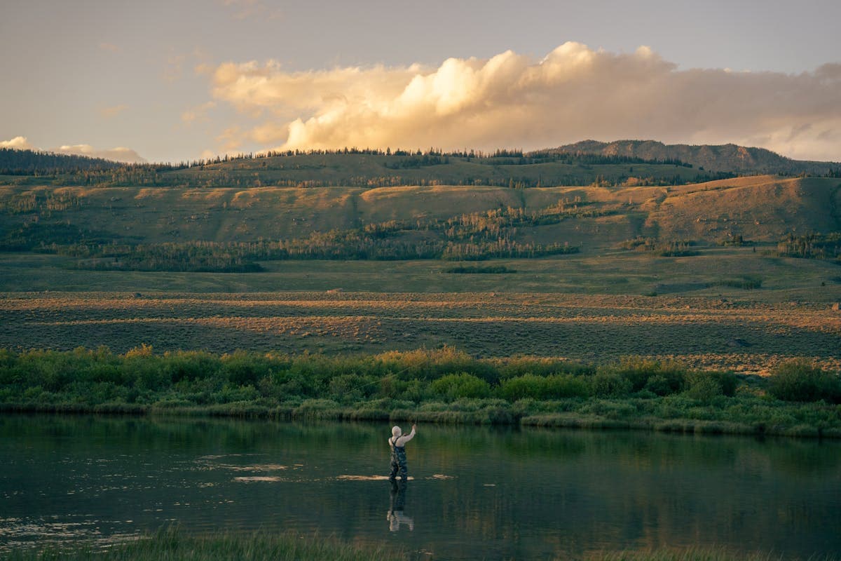 A lone fly angler casting on a wide flat Western river at dusk with sagebrush hills behind