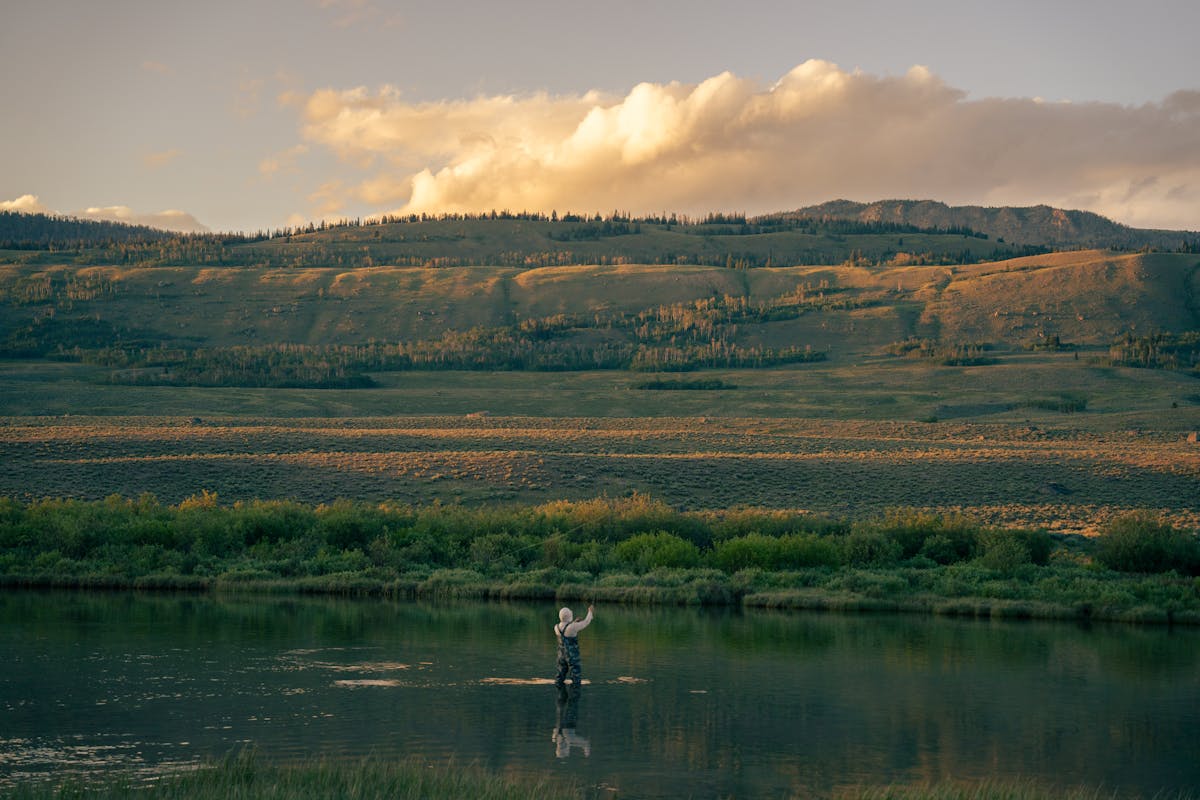 A lone fly angler casting on a wide flat Western river at dusk with sagebrush hills behind
